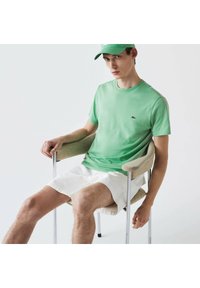 Lichtgroene katoenen t-shirt met een ronde hals en een klein logo op de borst, gecombineerd met witte shorts. Het model zit in een moderne stoel.