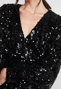 Kvinna klädd i ett svart plagg täckt av paljetter med djup V-ringning och textil som är glänsande och reflekterar ljus.