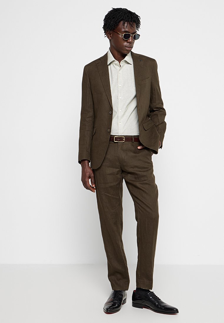 Brauner Leinenanzug mit einem hellgrauen Hemd, Gürtel und schwarzen Schuhen. Der Anzug hat eine maßgeschneiderte Passform und strukturierte Stoffe. Sonnenbrillen runden den Look ab.