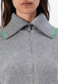 Šedý žebrovaný svetr s vysokým límcem, který má zelený akcent na límci a přední zip. Měkká struktura s dobře propracovanými designovými detaily.