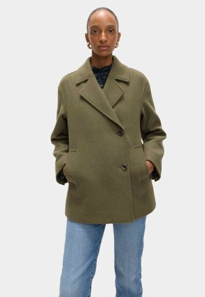Manteau en laine vert olive avec un design croisé, des revers crantés, deux poches latérales et de grands boutons. Associé à un jean bleu clair.