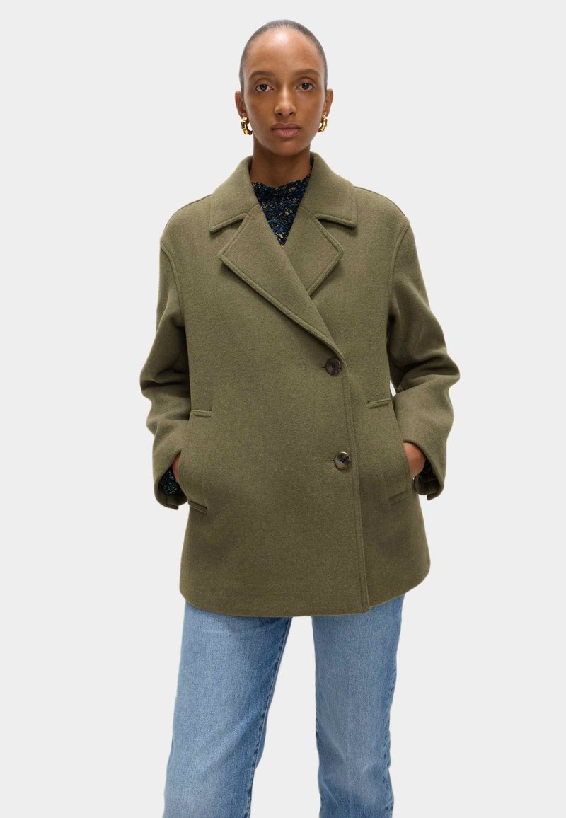 Olijfgroene wollen jas met een dubbelgeknopte ontwerpen, met geklapte revers, twee zijzakken en grote knopen. Gecombineerd met lichtblauwe jeans.