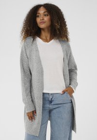 Szürke kötött kardigán nyitott elejű, bordázott mandzsettákkal és oldalsó hasítékokkal. Fehér V-nyakú pólóval és világoskék farmerral párosítva.