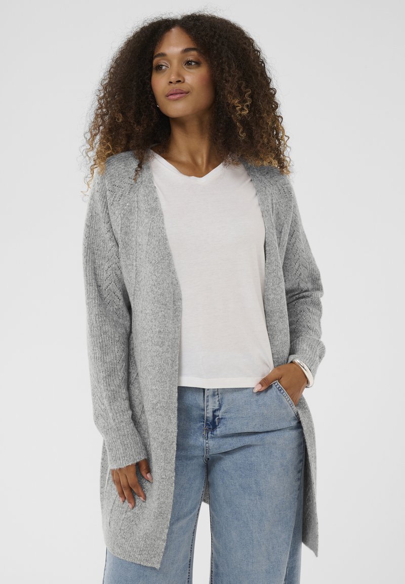 Szürke kötött kardigán nyitott elejű, bordázott mandzsettákkal és oldalsó hasítékokkal. Fehér V-nyakú pólóval és világoskék farmerral párosítva.