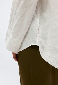 Λευκό λινό πουκάμισο με μακριά μανίκια, πλευρική σχισμή και μικρή ροζ και καφέ διακόσμηση με σταυροβελονιά, φορεμένο πάνω από χακί παντελόνια.