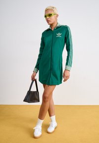 Vestito verde con zip, tre strisce bianche sulle maniche, polsini a costine e logo adidas. Abbinato a sneaker bianche e una borsa nera.