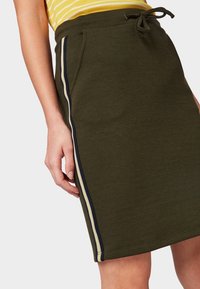 Olivově zelená sukně ke kolenům vyrobená z texturované tkaniny, s bočními kapsami a kontrastními námořnickými a bílými pruhy po stranách.