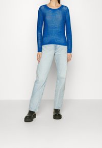 En person som bär en semitransparent ljusblå stickad tröja, ljus tvättade raka jeans och svarta kraftiga plattformskängor mot en neutral bakgrund.