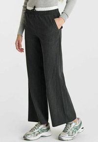 Pantalons à larges jambes à rayures noires avec une taille élastique et un cordon de serrage. Deux poches latérales, accompagnés de chaussures de sport argentées et vertes.