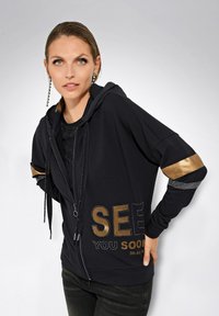 Schwarze Kapuzenjacke mit goldenen Akzenten, Reißverschluss und strukturiertem Schriftzug. Verfügt über eine lockere Passform und kontrastierende gemusterte Ärmel.