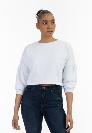 Felpa bianca corta con maniche lunghe e a sbuffo, abbinata a jeans blu scuro. Tessuto morbido e scollatura ampia; design minimalista.
