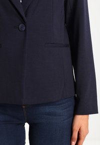 Námořnický modrý blazer s jedním knoflíkem, dvěma předními kapsami, přiléhavým střihem a hladkou texturou, v kombinaci s tmavými džíny.