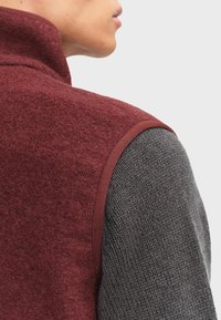 Burgunderfarvet uldsweater med høj krave, sat mod et mørkegrå tekstureret stof. Har en kontrasterende ribbet kant langs armhulen.