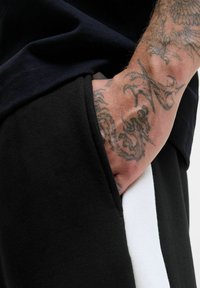 Sorte joggers med en hvid stribe, lavet af blødt stof. Hånden er placeret i sidelommen, hvilket fremviser detaljerede tatoveringer på armen.
