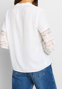 Blouse blanche avec des détails en dentelle sur les manches, présentant des rayures texturées et une coupe décontractée. Associée à un jean bleu, mettant en avant un style décontracté.