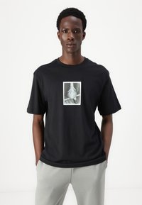 Sort bomulds t-shirt med et centralt grafik af en hånd, der holder en taske, korte ærmer og en afslappet pasform. Enkel design, ingen mønstre.