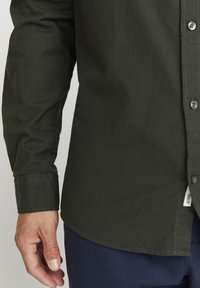 Camisa de botón verde oscura hecha de una tela texturizada; presenta un cuello, mangas largas y un dobladillo curvado. Se aprecian algunas costuras visibles cerca de los puños.