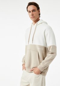 Beige en witte color-blocked hoodie met een voorzak, een capuchon met trekkoord en een gladde textuur. Bevat minimale tekst in een subtiele print.