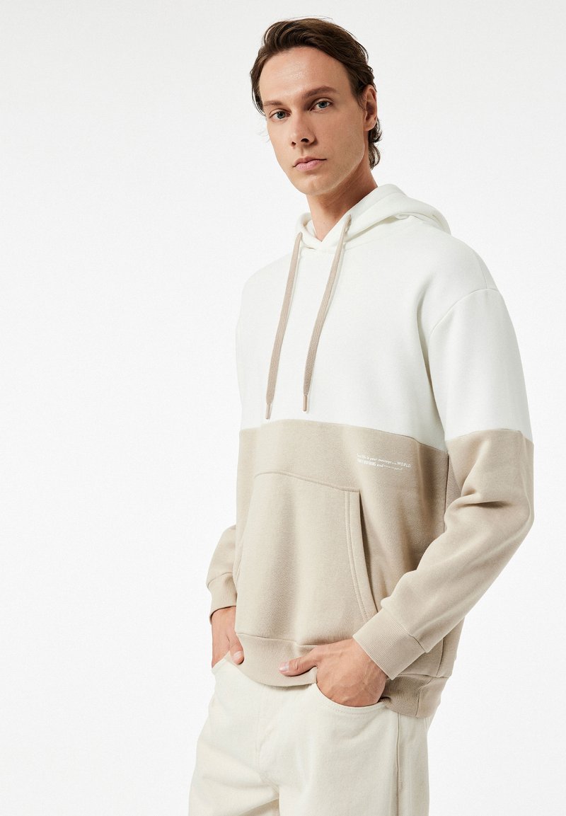 Beige en witte color-blocked hoodie met een voorzak, een capuchon met trekkoord en een gladde textuur. Bevat minimale tekst in een subtiele print.