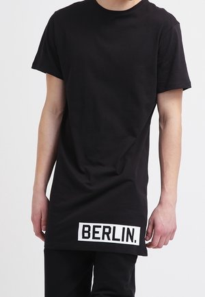 Person iført en lang sort T-shirt med "BERLIN." trykt med fede hvide bogstaver nær den nederste kant.
