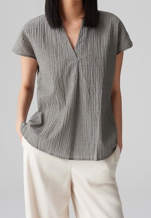 Personne portant un haut gris à manches courtes texturé avec un col en V et un pantalon blanc ample, debout les mains dans les poches devant un fond gris.