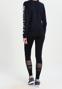 Felpa navy con logo sulla manica, abbinata a leggings neri con dettagli in rete. Le scarpe sono navy con suole bianche.