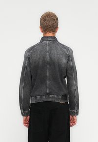 Persona con cabello corto rubio vistiendo una chaqueta de mezclilla negra desteñida y pantalones negros, de pie frente a un fondo blanco liso.