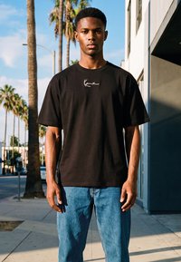 Joven con camiseta negra con un logo de firma blanca y vaqueros azules, de pie en una acera urbana iluminada por el sol, con palmeras.