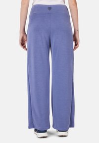 Lila weit geschnittene Hose aus weichem Stoff, mit hohem Bund und einer Logo-Applikation auf der Rückseite, kombiniert mit weißen Sneakern mit Farbspritzern.