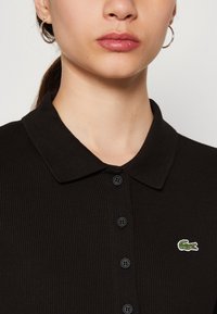 Polo shirt nera a coste con colletto a punta, cinque bottoni e un piccolo logo di coccodrillo ricamato in verde sul lato sinistro del petto.