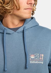 Blue hoodie with drawstrings, featuring a graphic print with tropical motifs and the text "Billabong" in white. Soft texture and casual design.