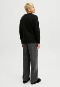 Berniukas su trumpais šviesiais plaukais, vilkintis juodą megztinį, pilkas plačias kelnes ir juodus batus, stovi šonu prieš paprastą foną.