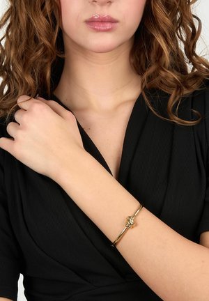 Donna con capelli ricci che indossa un top nero e un braccialetto nodo dorato al polso sinistro, mano appoggiata sulla spalla destra.