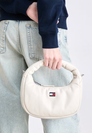 Osoba trzyma małą białą torebkę z wyściełaną rączką i logo Tommy Jeans, ubrana w jasnoniebieskie dżinsy i ciemnoniebieski top z długim rękawem.