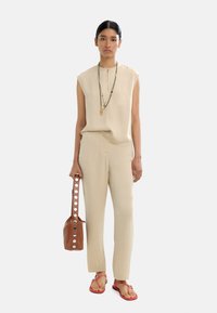 Beżowy top bez rękawów i dopasowane spodnie, brązowa torba z okrągłymi akcentami oraz czerwone płaskie sandały. Gładka faktura materiału z prostym wzorem.