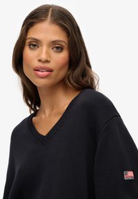 Granatowy sweter dzianinowy z dekoltem w serek, z ribbrowym wykończeniem i teksturowanym wykończeniem. Zawiera naszywkę z akcentem amerykańskiej flagi.