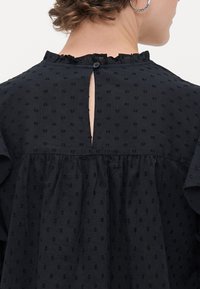 Blusa nera con scollatura arricciata e chiusura con bottoni sul retro. Presenta un tessuto testurizzato con un leggero motivo a pois e una vestibilità morbida.