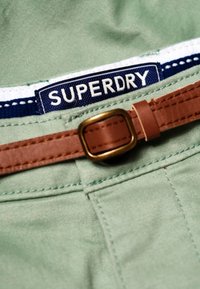 Cinturilla de tela verde con cinturón de cuero marrón que presenta una hebilla dorada. Etiqueta azul con texto blanco "SUPERDRY" visible en la cinturilla.