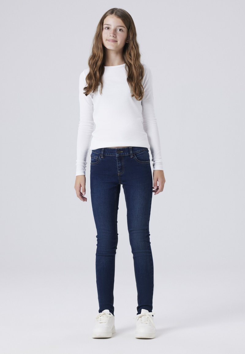 Camisa blanca de manga larga, jeans ajustados azul oscuro y zapatillas blancas. Diseño simple con tejido suave y detalles mínimos.