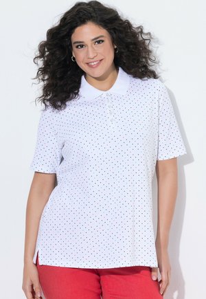 Uśmiechnięta kobieta o kręconych włosach, ubrana w białą koszulkę polo z krótkim rękawem z małymi kolorowymi kropkami i czerwone spodnie na białym tle.