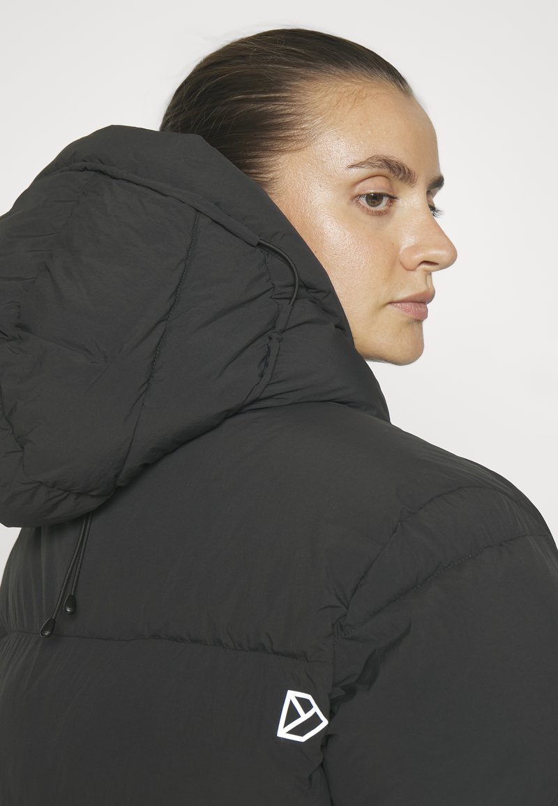 Frau mit nach hinten gekämmtem Haar, die eine schwarze Daunenjacke mit Kapuze trägt, von hinten betrachtet und nach rechts schauend.