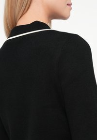 Melnā adīta kardigāna ar strukturētu apdari, kuram ir balta svītru akcentācija gar apkakli. Dizainā iekļautas noapaļotas malas un pieguļošs siluets.