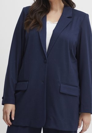 Mujer con una chaqueta azul oscuro sobre una camiseta blanca, con el cabello largo y oscuro, mostrado desde los hombros hasta las caderas contra un fondo liso.
