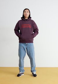 Bordo spalvos džemperis su raudonu tekstu, priekinė kišenė, derinamas su šviesiai mėlynomis kelnėmis. Modelis stovi ant geltono grindų fono priešais paprastą baltą foną.