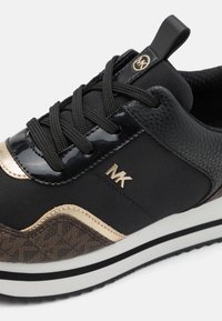 Zwarte sportieve sneaker met leren, mesh en gemusterde accenten. Heeft een gouden logo, dikke witte zool en zwarte veters met een gevoerde kraag.