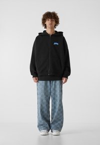 Sudadera negra con cremallera y un logo azul, combinada con pantalones sueltos de patrón azul y zapatillas blancas. Diseño simple y estilo casual.