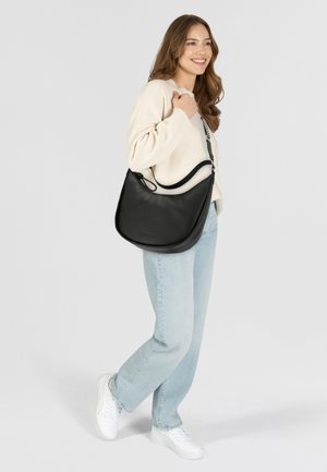 Borsa a spalla in pelle nera con design curvo, tracolla regolabile e dettagli in metallo argentato, tenuta da una persona in abbigliamento casual.