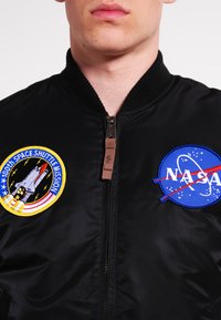 Černá bomber bunda s žebrovaným límcem, zdobená dvěma vyšívanými nášivkami: logem NASA a designem 100. mise Space Shuttle. Viditelný detail zipu.