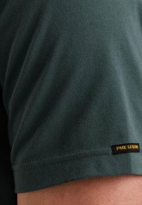 Tamnozeleni majica kratkih rukava iz mekog materijala, s PME Legend logotipom u žutoj boji na rubu. Gladak materijal, minimalistički dizajn.