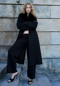 Abrigo negro con cuello de pelo sintético, combinado con pantalones negros holgados y tacones abiertos negros. Pared de piedra texturizada en el fondo.